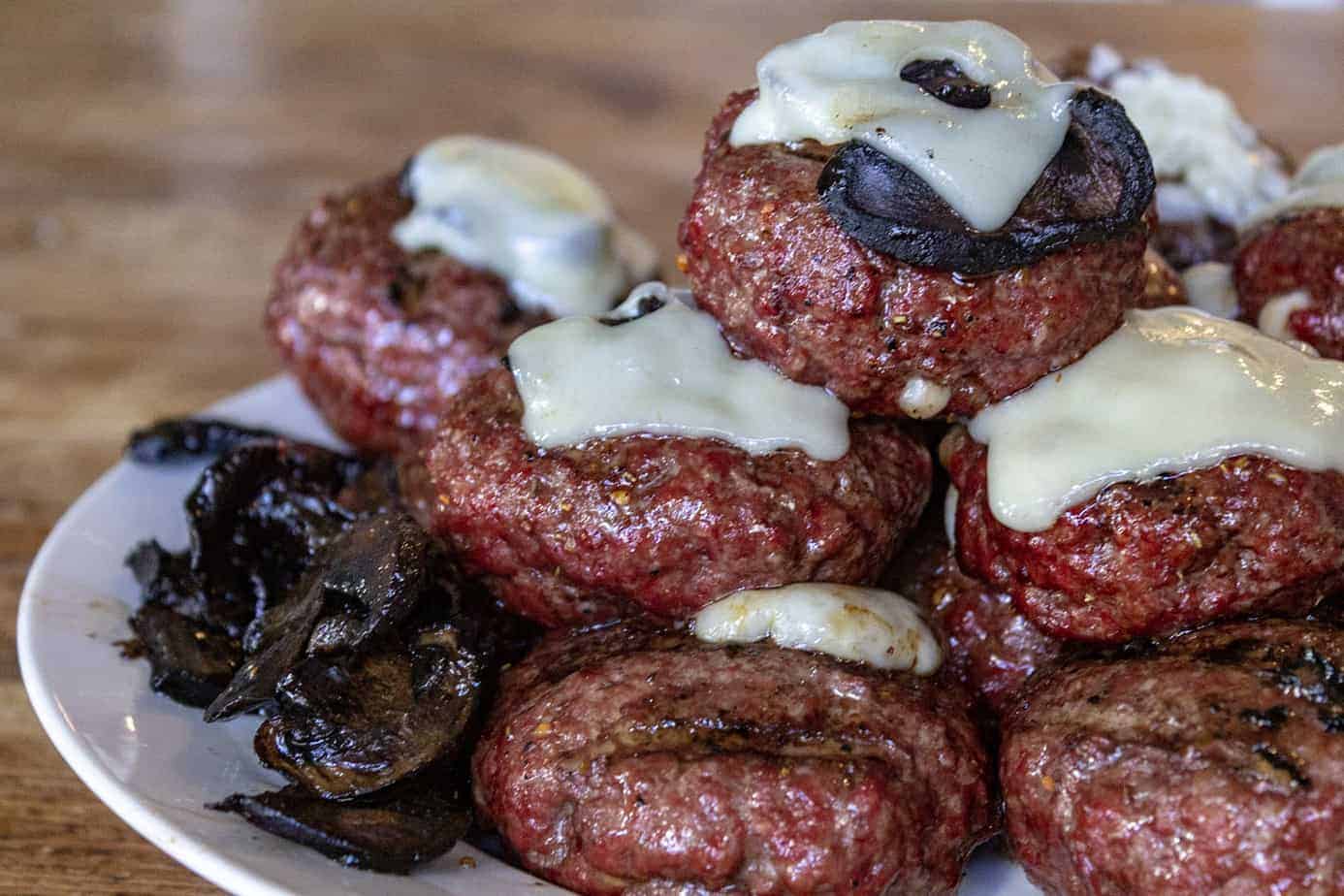 A pile of smoked juicy lucy hamburgers topped with provolone cheese and sitting next to some cooked mushrooms.