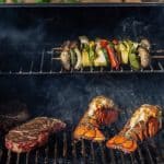 Dry aged ribeyes, vegetable kabobs, and lobster tails on a grill. Smoke can be seen lightly swirling around the food.