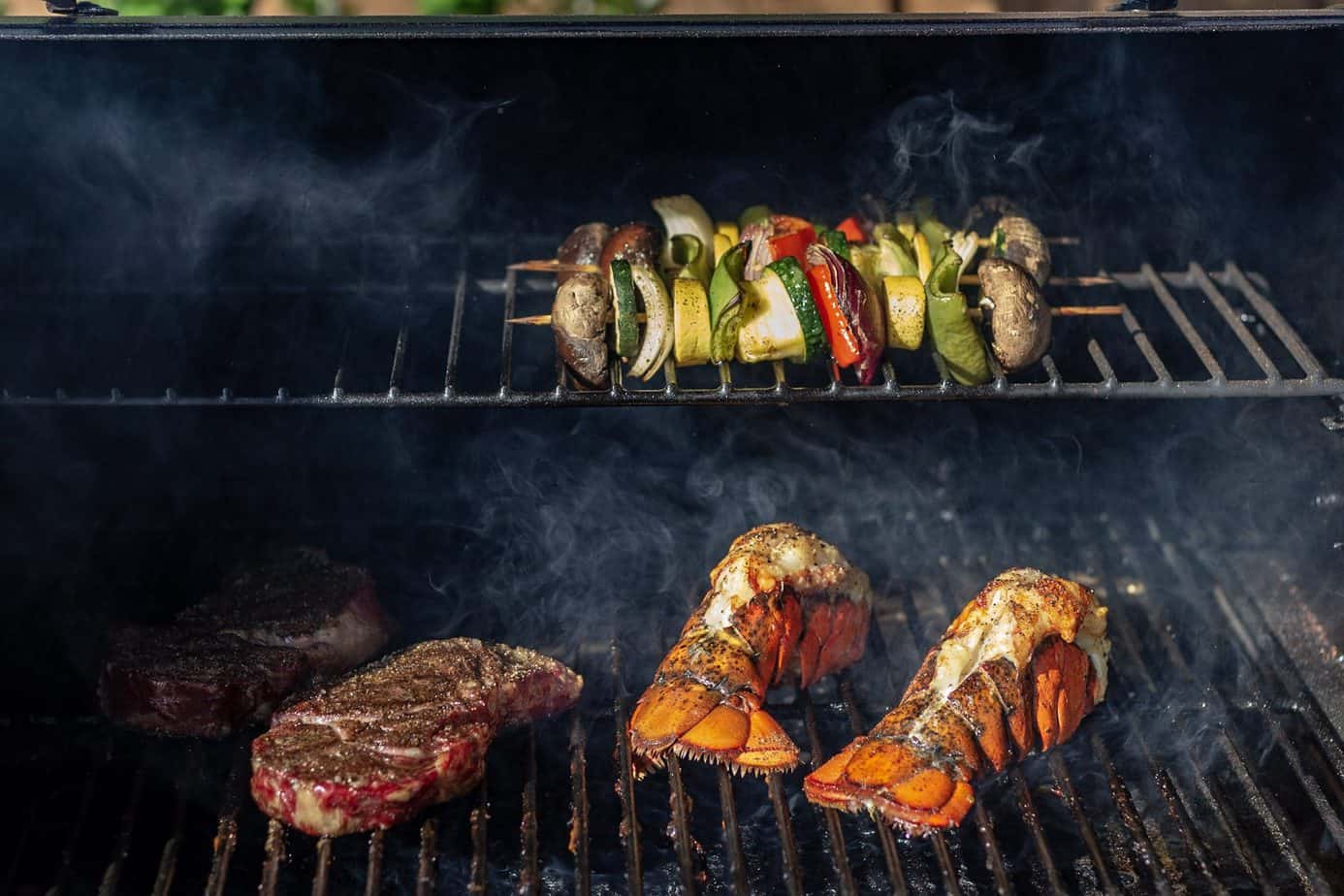 Dry aged ribeyes, vegetable kabobs, and lobster tails on a grill. Smoke can be seen lightly swirling around the food.