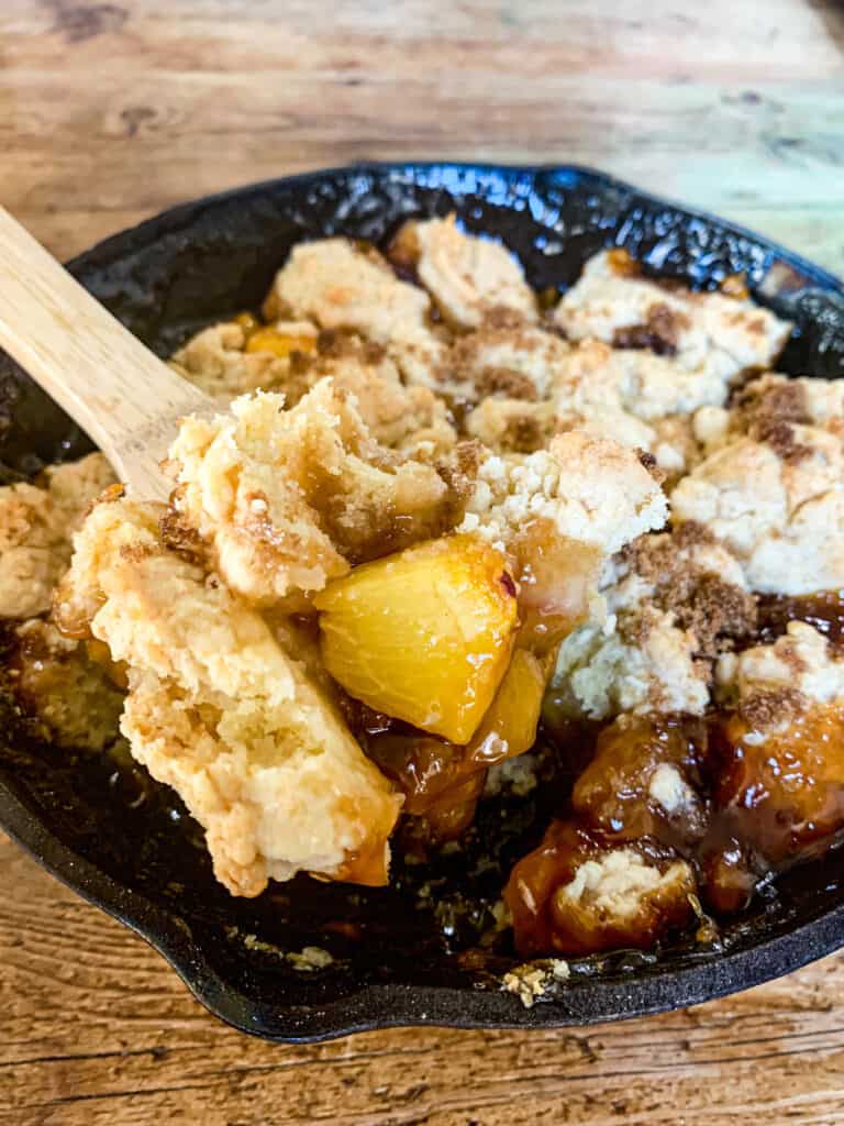 A scoop of peach cobbler on a spoon over a cast iron skillet